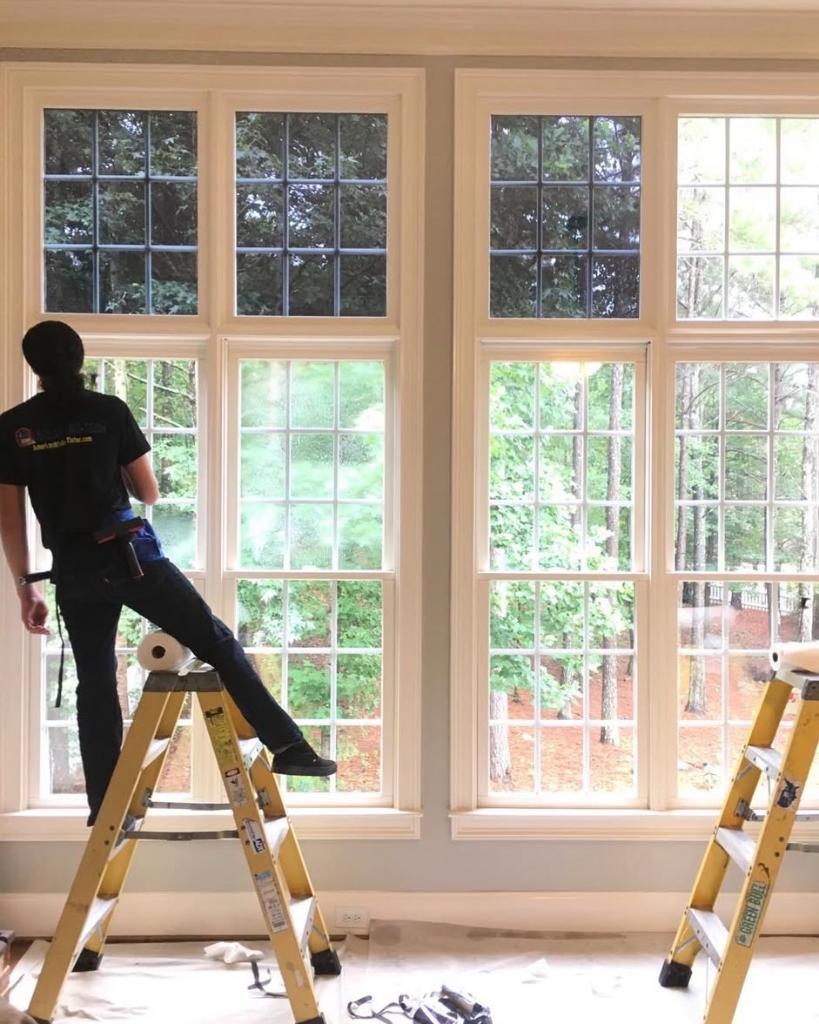 Technician on ladder applying window film to large residential windows with forest view